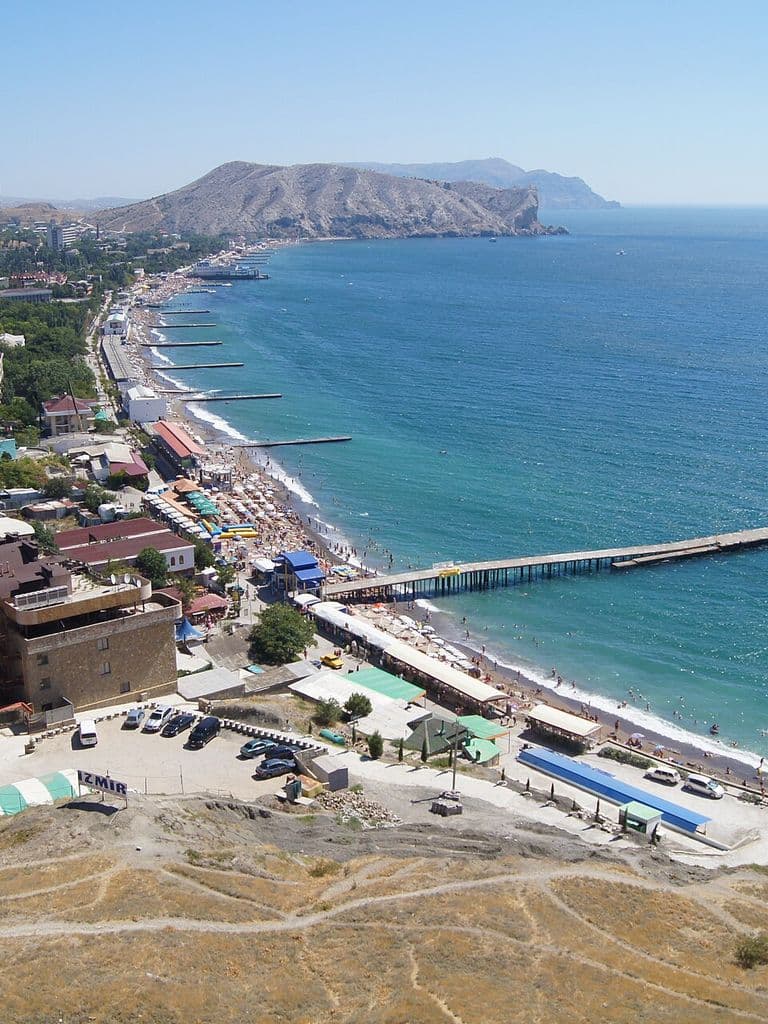 Судакская бухта с высоты: дуга пляжа и набережная Судака, пирс в море, вдали мыс Алчак