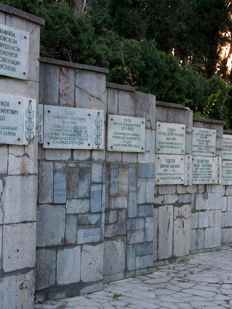 Памятная стена Поликуровского мемориала в Ялте с плитами в честь похороненных на бывшем Массандровском кладбище, за ней кипарисы в утреннем свете