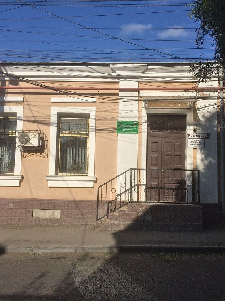 Одноэтажное здание музея крымчаков на улице Крылова в Симферополе: светлый фасад с зарешёченными окнами, высоким крыльцом и воротами