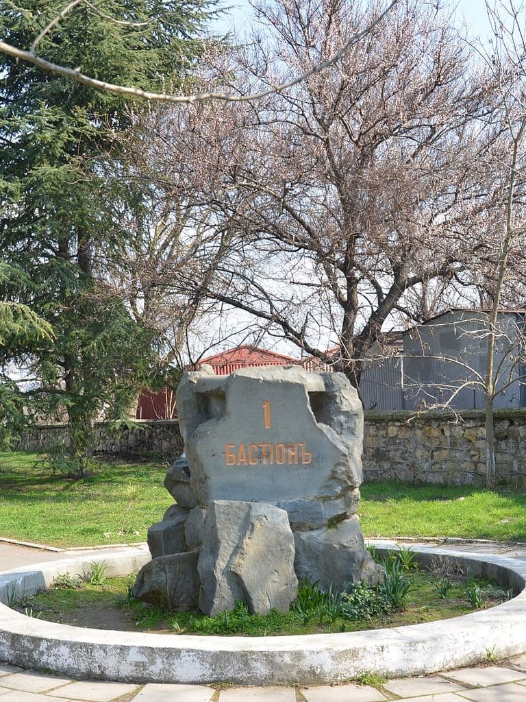 Памятник Первому бастиону в Севастополе: глыба тёмного камня с надписью в круглой клумбе, вокруг сквер с кедрами и мощёные дорожки